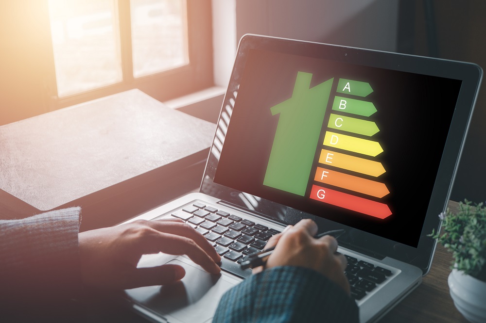 Człowiek siedzi przy laptopie (widok na laptop i dłonie na klawiaturze). Na ekranie wyświetla się ikona domu połączona z paskami symbolizującymi klasę energetyczną
