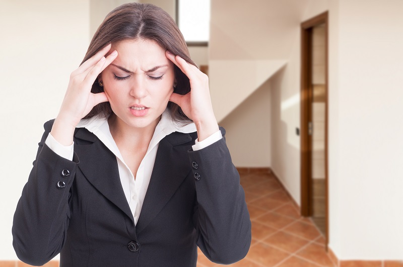 Młoda kobieta w stroju biznesowych stoi w pustym mieszkaniu i trzyma się za głowę, z grymasem napięcia lub bólu